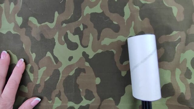 Close-up hand removes cat hair, threads, litter with roller lint brush from militar patterned fabric