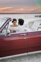 Beautiful couple at sunset on a summer wedding day. Romantic couple in love next to their convertible. The groom is in a light suit, the bride is in a beautiful stylish dress.