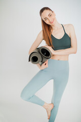A young woman yoga instructor stands in a tree pose, smiles and holds mats sunbeam shine on her body