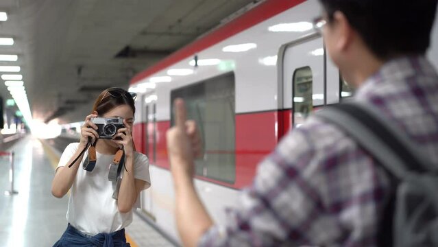 Selective focus of a happy beautiful Asian female traveler standing on a subway platform next to a train, using a vintage film camera taking a picture of a blurred Asian male traveler with backpack.