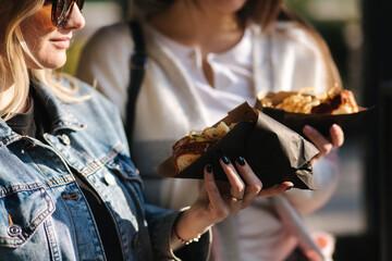 Tasty hot-dog in female's hands. Girls buy street food. Two beautiful blonde and brunette girls...