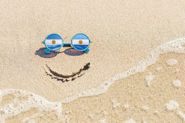 Fototapeta na wymiar A painted smile on the sand and sunglasses with the flag of Argentina. The concept of a positive and successful holiday in the resort of Argentina.