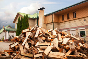 A pile of split firewood for heating the house, unloaded in the yard, against the backdrop of the house, natural heating sources.