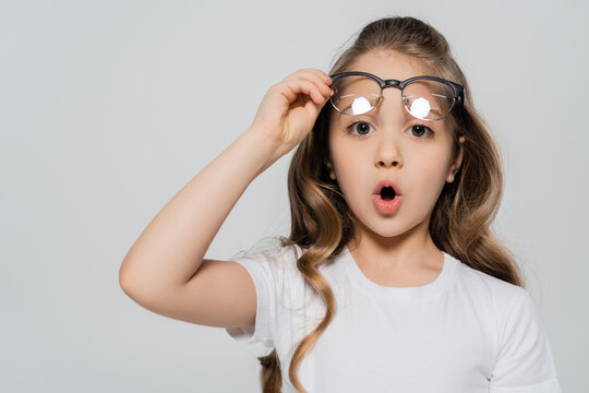 surprised girl with open mouth holding eyeglasses and looking at camera isolated on grey.
