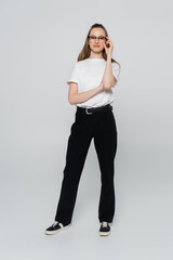 full length view of woman in white t-shirt and black trousers adjusting eyeglasses on grey background.