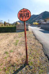 Heavily rusted fire hydrant metal display sign written in Japanese and english language standing not straight on dried grass aside an asphalt countryside road with mountains and houses in background.