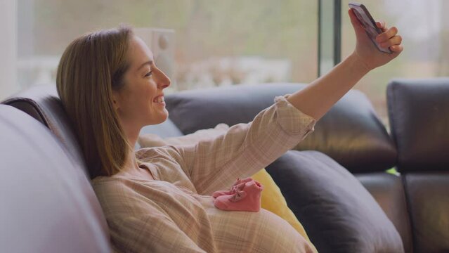 Pregnant woman sitting on sofa at home taking selfie of small baby shoes  on stomach with mobile phone - shot in slow motion