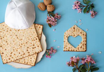 Layout of Matzah shape of small house with heart,  matzo on plate, pink flowers and white kipa on blue background. Traditional of Jewish Holiday on Passover. Home symbol of happy family. Top view. 