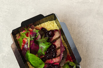 Healthy box with turkey with cherry, greens and couscous in eco paper black takeaway container on gray background. Restaurant food delivery concept.
