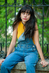 Young girl dressed in yellow and wearing a denim jumpsuit sitting on a stone wall in a park with autumn green and yellow colors