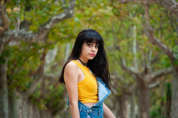 Young girl dressed in yellow and wearing a denim overalls in profile with a park background full of trees in autumn shades of green and yellow