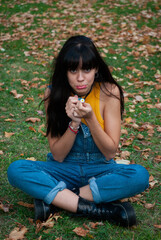 Fototapeta premium Young girl dressed in yellow and wearing a denim overalls sitting while playing sadly with a daisy in a park of autumn green and yellow colors