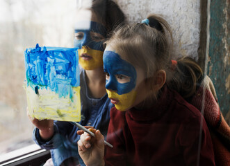 Two little girls with Ukrainian flags painted on their faces draw a picture on the glass. Children against war.