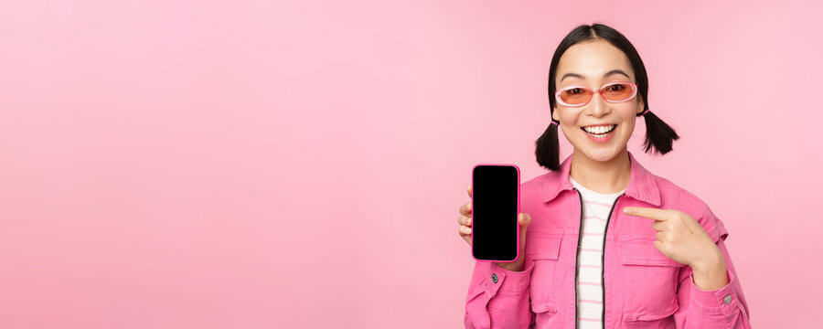 Enthusiastic Asian Woman In Stylish Clothes, Sunglasses, Pointing Finger At Mobile Phone Screen, Showing Smartphone Application, Standing Over Pink Background