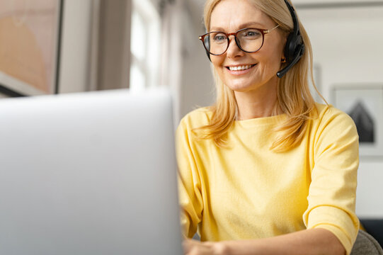 Online consultant in touch. A middle-aged woman wearing headset talking on the distance with a customers, clients sitting at the desk with a laptop in a modern office space