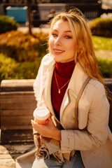 Obraz premium a beautiful blonde with a cup of coffee sits on a bench in an autumn park.