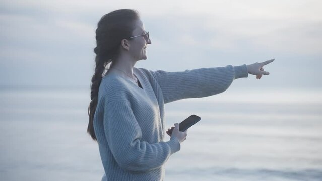 Young beautiful woman points to sea and shoots photo or video on smartphone. Brunette caucasian adult female digital nomad with braid in sunglasses and loose grey sweater enjoys seaside view.