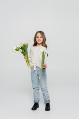 full length view of girl in jeans and blouse showing white tulip at camera on grey background.