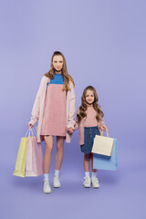 full length of pleased mother and daughter holding shopping bags on purple.