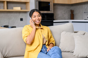 Obraz premium Happy African-American woman talks via smartphone sitting on the sofa at home, charming lady enjoys phone conversation staying at home, glad to talk with friend