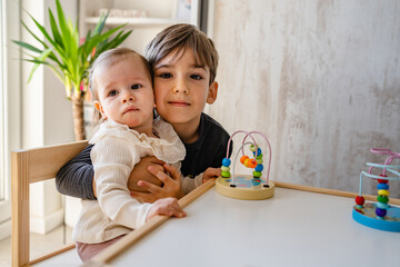 One small caucasian boy five years old holding his little baby sister girl while playing at home in room in bright day front view copy space family siblings love concept looking to the camera