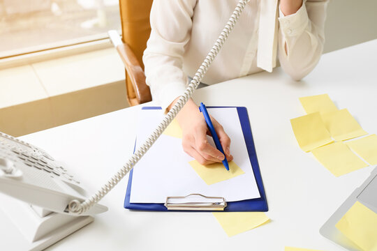 Young manager writing on sticky note paper while talking by phone at her workplace