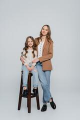 trendy woman embracing daughter sitting on high stool on grey background.