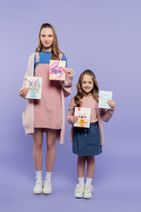 full length of mother and happy daughter holding greeting cards on purple, mothers day concept.