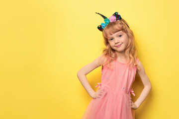 Close-up portrait of a cute attractive cheerful girl. Cute smiling child wearing a festive unicorn headband. Blonde girl 5 years old in a pink dress on a bright yellow background looking at the camera