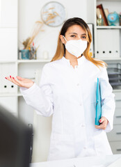 Fototapeta na wymiar Portrait of young hispanic female doctor in face mask making welcome gesture welcoming patient to clinic