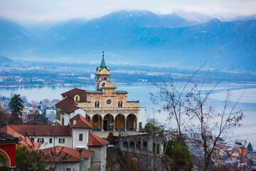 View of principal sight and goal of pilgrimage in Switzerland, Madonna del Sasso church on sacred mountain on background of Lake Maggiore and Locarno cityscape in misty winter day