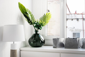 Corner of a room furnished with a white wooden chest of drawers, under an aluminum window with decorative plants and a matching lampshade lamp