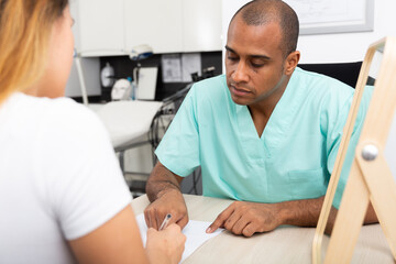 Experienced Hispanic doctor cosmetologist working with female client in clinic, filling out medical form before procedure