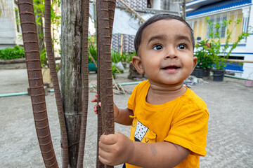 A infant child is trying to stand up holding a rusty iron rod in his small two hands. A kid is having fun and sticking out his tongue.