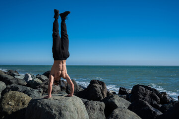 Shirtless man doing handstand on rocks by the sea. 