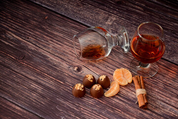 Two glasses of cognac, chocolates, mandarin slices and cinnamon sticks on a wooden table.