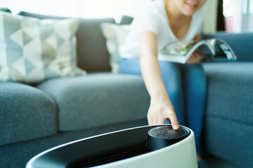 Happy Asian young woman turning on high efficiency air purifier while staying and reading a book in...