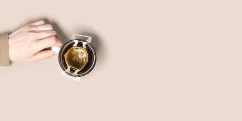 Woman hand hold cup and make black coffee drink in cup, drip coffee bag with ground coffee brewed in cup, paper bag drip coffee filter, beige background, top view, minimal flat lay