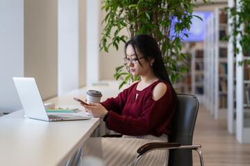 Young asian student girl procrastinate in library using smartphone and chatting in social media while preparing for exam test. Lazy woman with mobile phone in hand avoid learning and doing homework