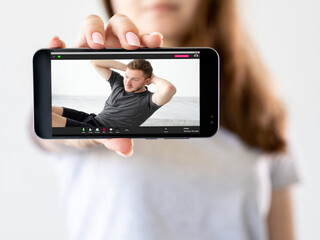 Virtual fitness class. Online training. Workout tutorial. Girl hand showing sport video program on phone with athletic man doing sit-ups on screen isolated on defocused neutral.