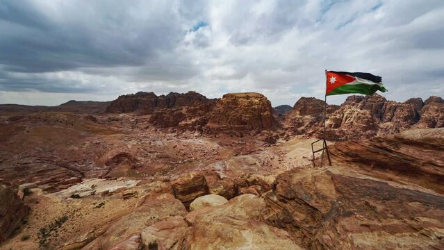Cinemagraph seamless video loop of the Jordanian flag in Petra, Jordan, moving in the wind at a bedouin camp with vibrant colors and a view of the desert rocks with tombs carved into sandstone rock.