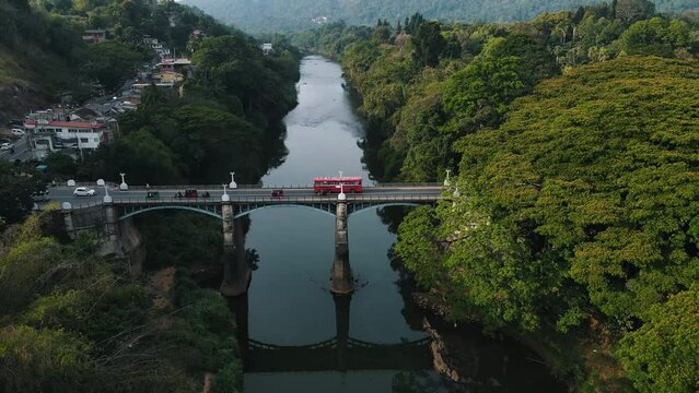 Several traditional Tuktuks ride between cars and buses over the tall historic Peradeniya Bridge over the reflected Mahaweli Ganga River in Sri-Lanka on a sunny spring day. Drone Dolly shot