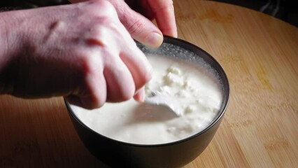 A close up shot of a black mixing bowl on a wooden kitchen table filled with white low fat cheese as a chef carefully breaks up clumps with the back of a spoon and stirring to create an even texture