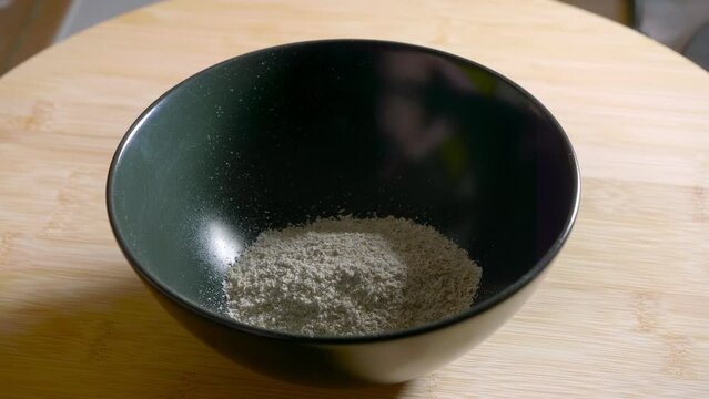 A close up of an empty black bowl on a wooden kitchen table as raw ground oatmeal, whey protein, low fat cheese and water is added to the bowl and is the ingredients for a low-calorie protein dessert