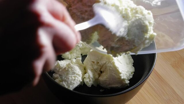 A close up shot of a black mixing bowl on a wooden kitchen table, a chef carefully preparing white low fat cheese by transferring it from a storage container and placing it into the bowl