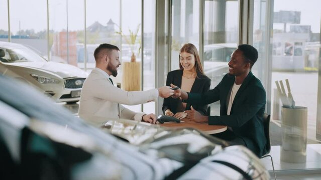 Happy multicultural couple sitting with salesman at auto salon and getting keys from their new car. Smiling man and woman doing payment with credit card. Couple sitting making purchase at auto salon.
