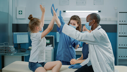 Cheerful nurse and medic giving highfive to small child after successful checkup appointment. Woman and specialist feeling happy at medical examination with girl and mother. Diverse team - obrazy, fototapety, plakaty