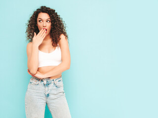 Sexy young beautiful smiling female in trendy summer hipster clothes. carefree woman posing near blue wall in studio. Positive brunette model with afro curls hairstyle. Shocked and surprised