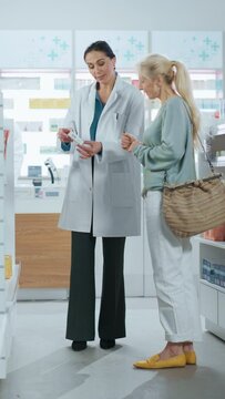 Vertical Video. Pharmacy: Professional Caucasian Female Pharmacist Helping Caucasian Senior Female Customer with Medicine Recommendation, Advice, Talking. Drugstore with Full of Health Care Products