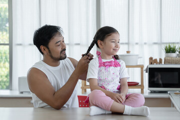 Happy Asian young father with beard making braid for his Caucasian little daughter while sitting on...